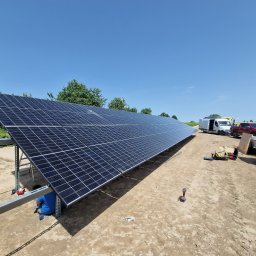 Usługi elektryczne F.W. - Montaż paneli słonecznych na konstrukcji stalowej w terenie wiejskim, widoczny pracownik w niebieskiej koszulce, samochody dostawcze i ciągnik w tle.