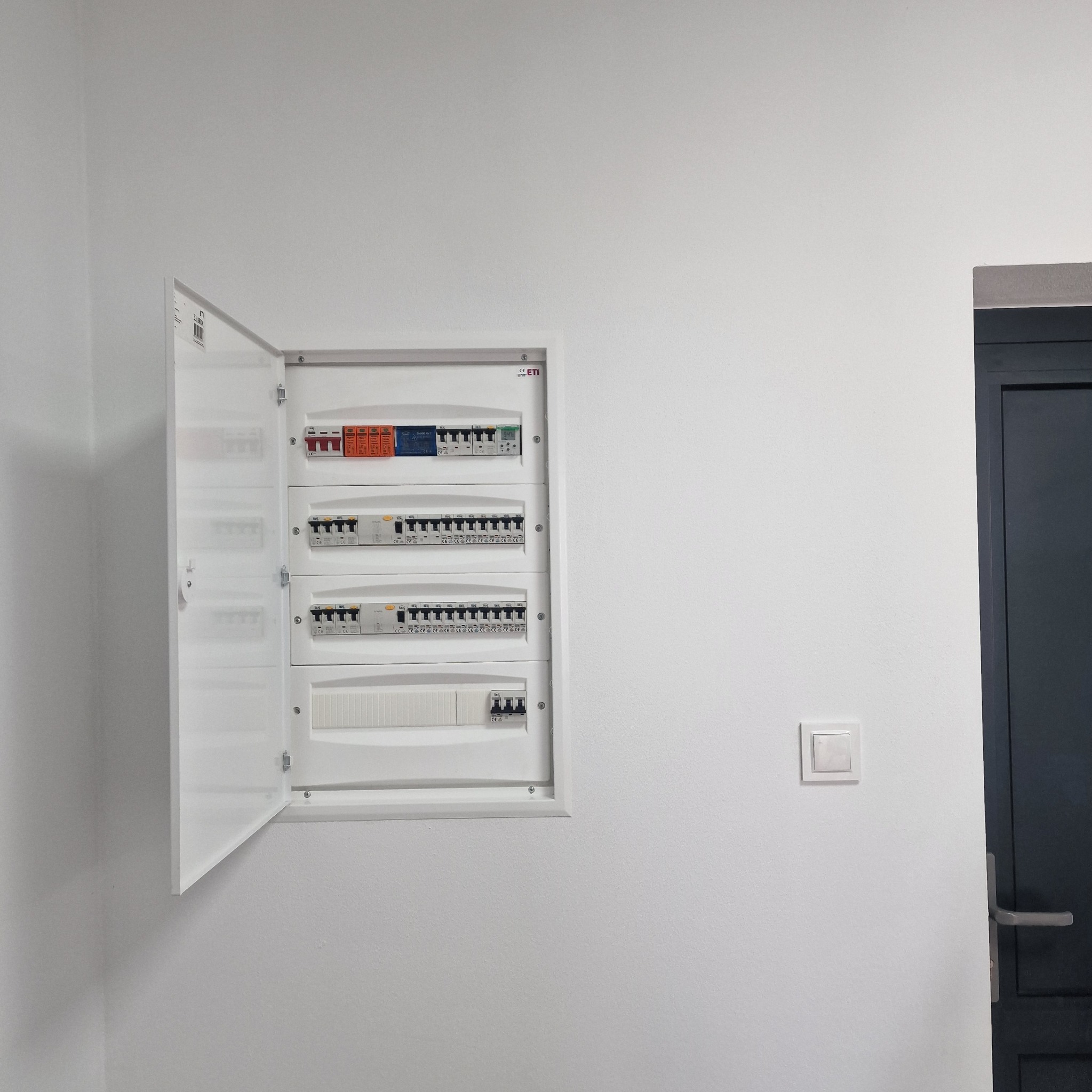 Otwarta biała skrzynka rozdzielcza z bezpiecznikami na białej ścianie, obok włącznik światła i fragment ciemnych drzwi.