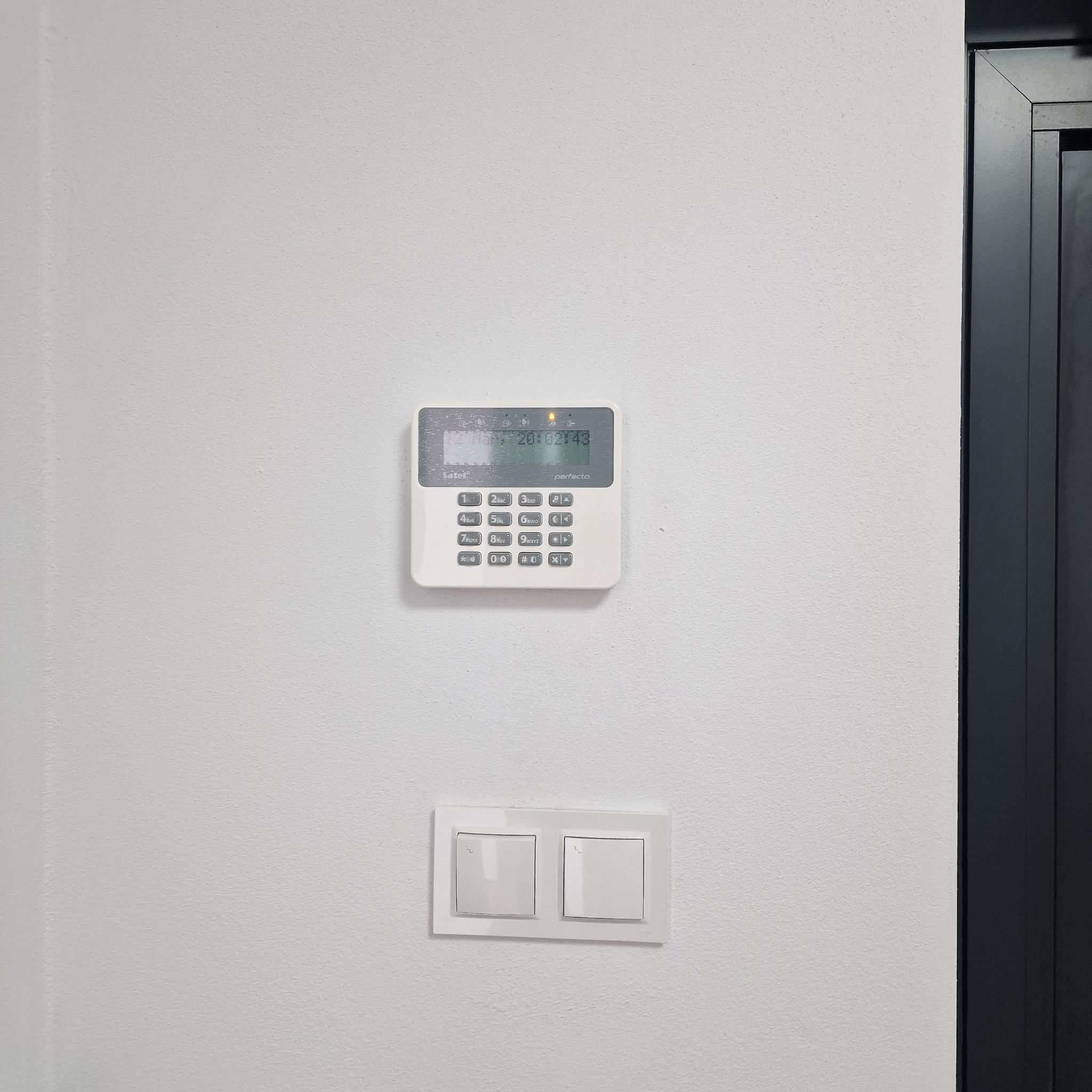 Biały panel sterowania alarmem domowym marki Satel, model Perfecta, zamontowany na białej ścianie obok włącznika światła i czarnej futryny drzwi. Na wyświetlaczu widoczny czas i status.