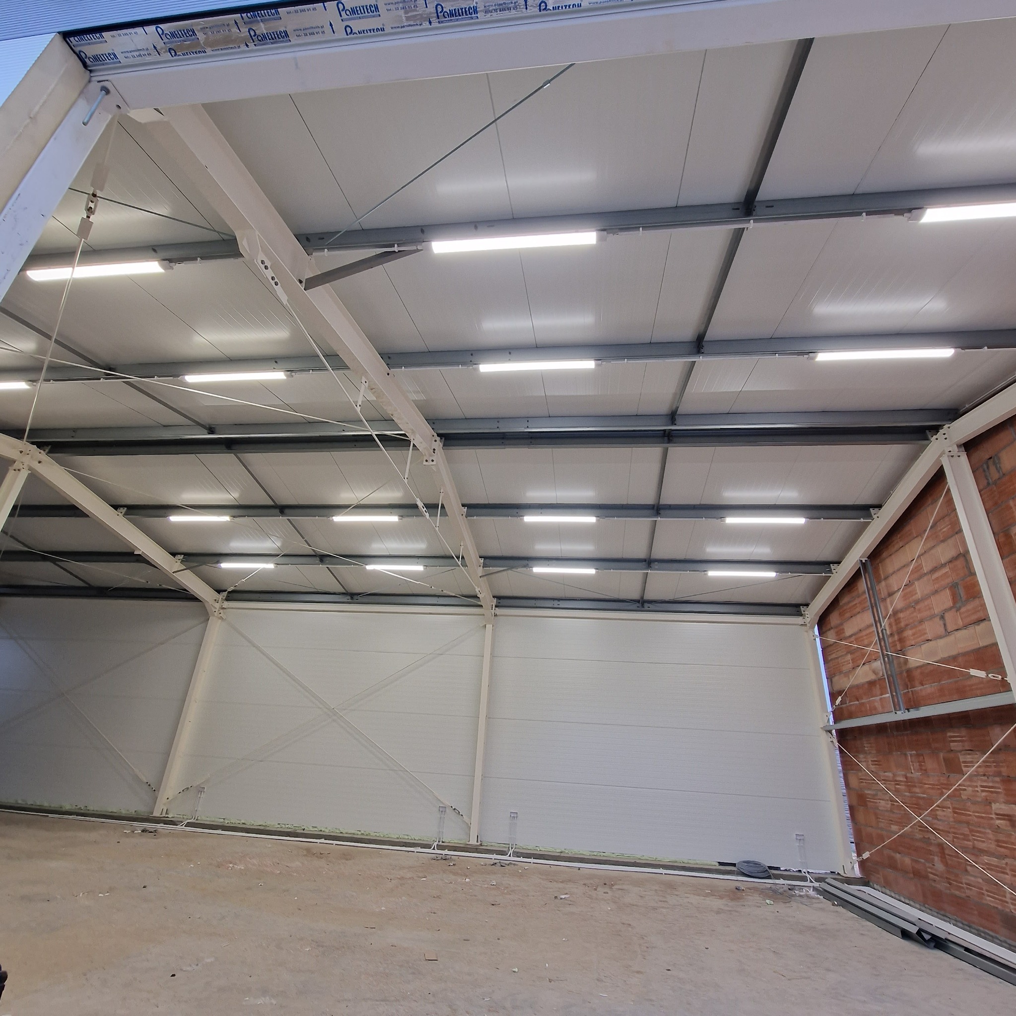 Wnętrze nowo wybudowanej hali z widoczną konstrukcją stalową, oświetleniem LED i częściowo wykończonymi ścianami z paneli warstwowych. Widoczny fragment muru z cegły.