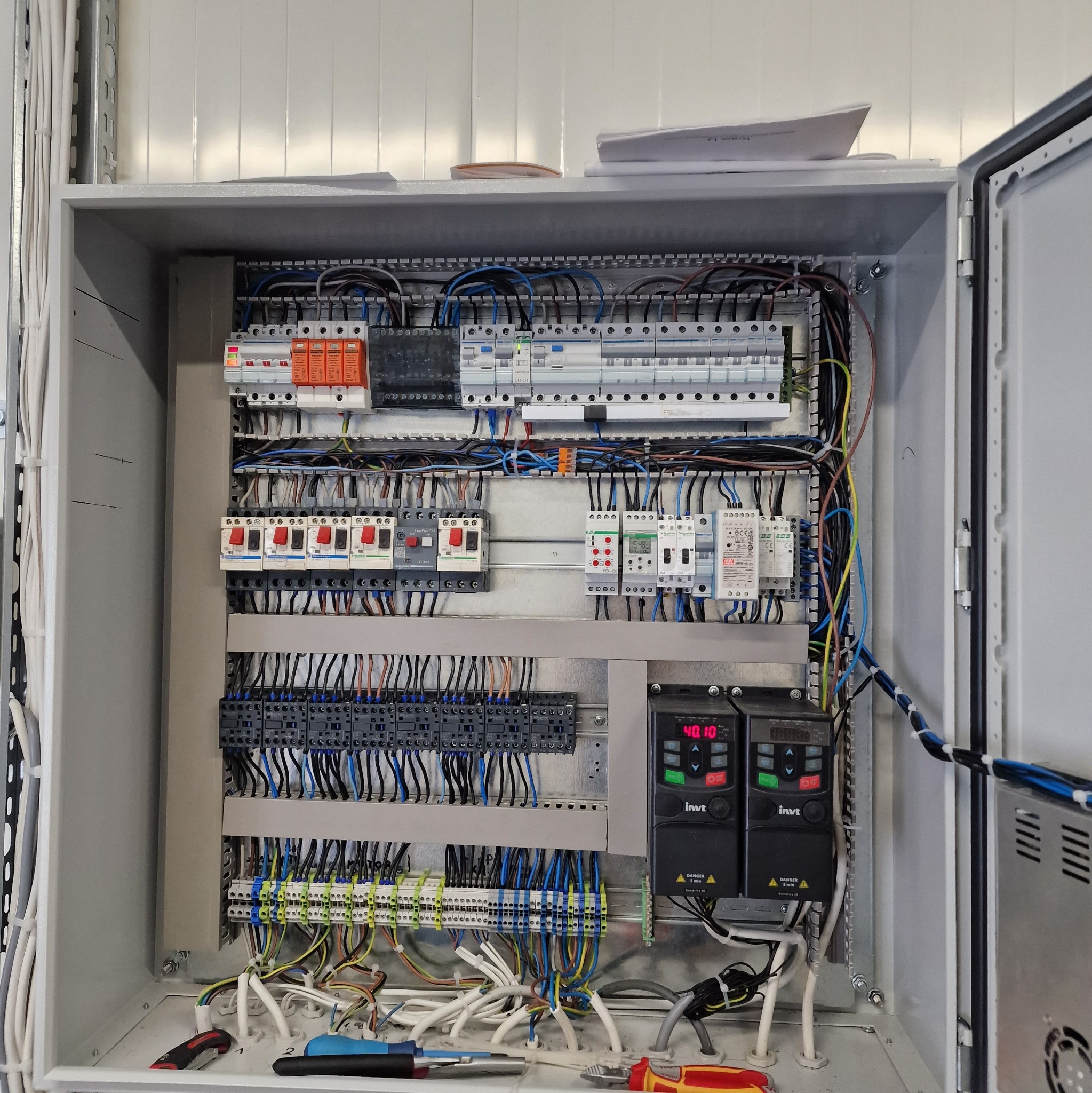 Otwarta szafa sterownicza w kolorze szarym, z widocznymi zabezpieczeniami, bezpiecznikami, przekaźnikami, sterownikami i licznymi przewodami; na dnie szafy leżą narzędzia - kombinerki i śrubokręt.