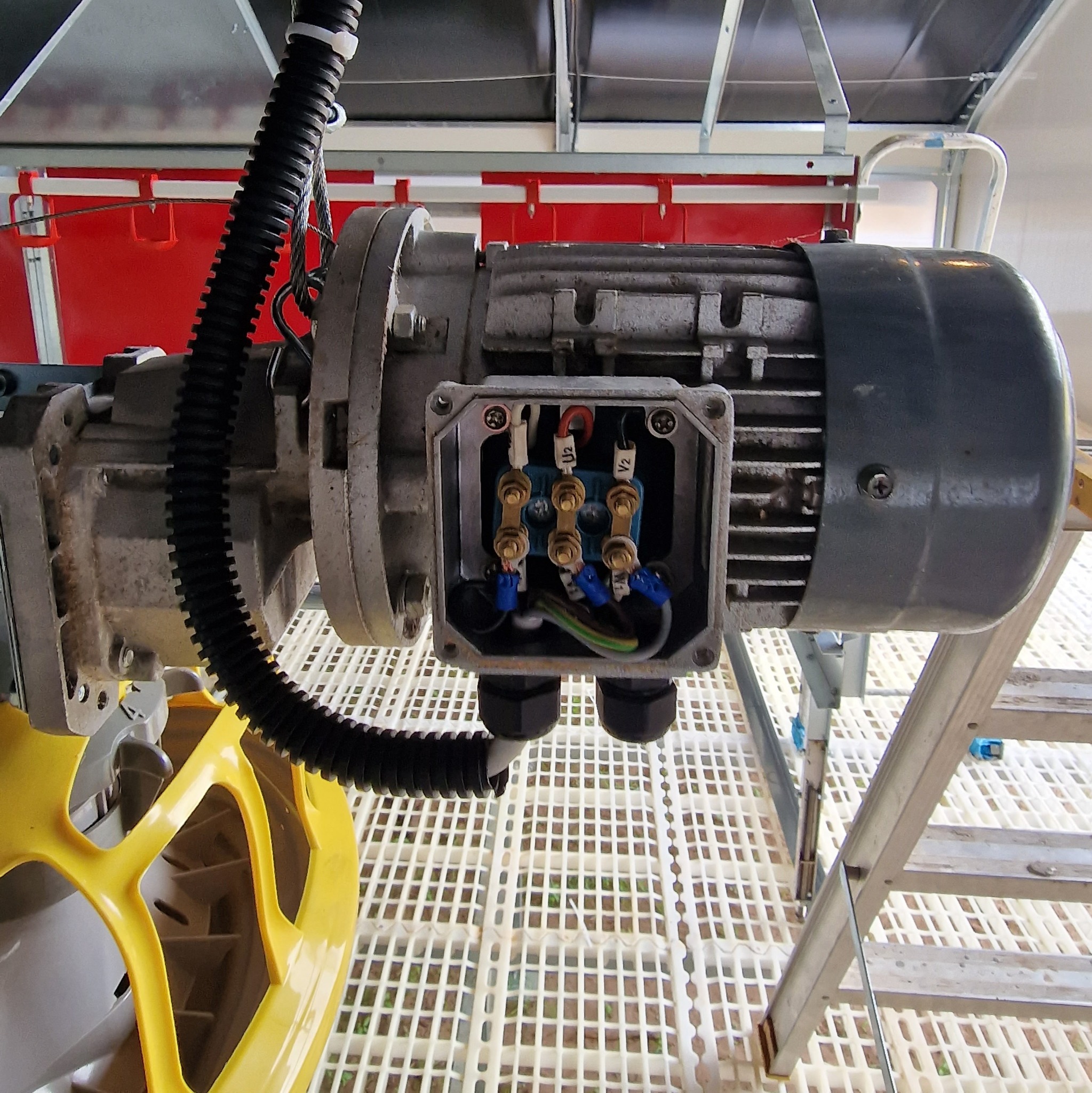 Szary silnik elektryczny z otwartą puszką przyłączeniową, widoczne przewody i złączki, czarna karbowana rura ochronna, żółty element konstrukcyjny i drabina w tle.