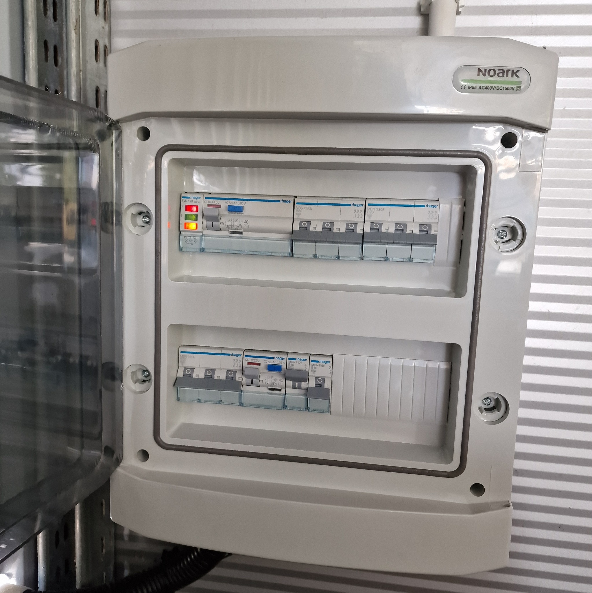 Otwarta skrzynka elektryczna marki Noark z widocznymi bezpiecznikami Hager, zamontowana na metalowym stelażu, z sygnalizacją LED na jednym z bezpieczników.
