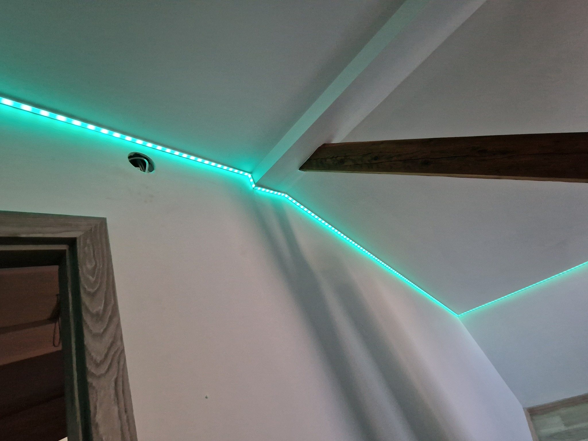 Turkusowe taśmy LED oświetlające sufit w rogu pokoju, widoczna drewniana belka i fragment ościeżnicy drzwi.