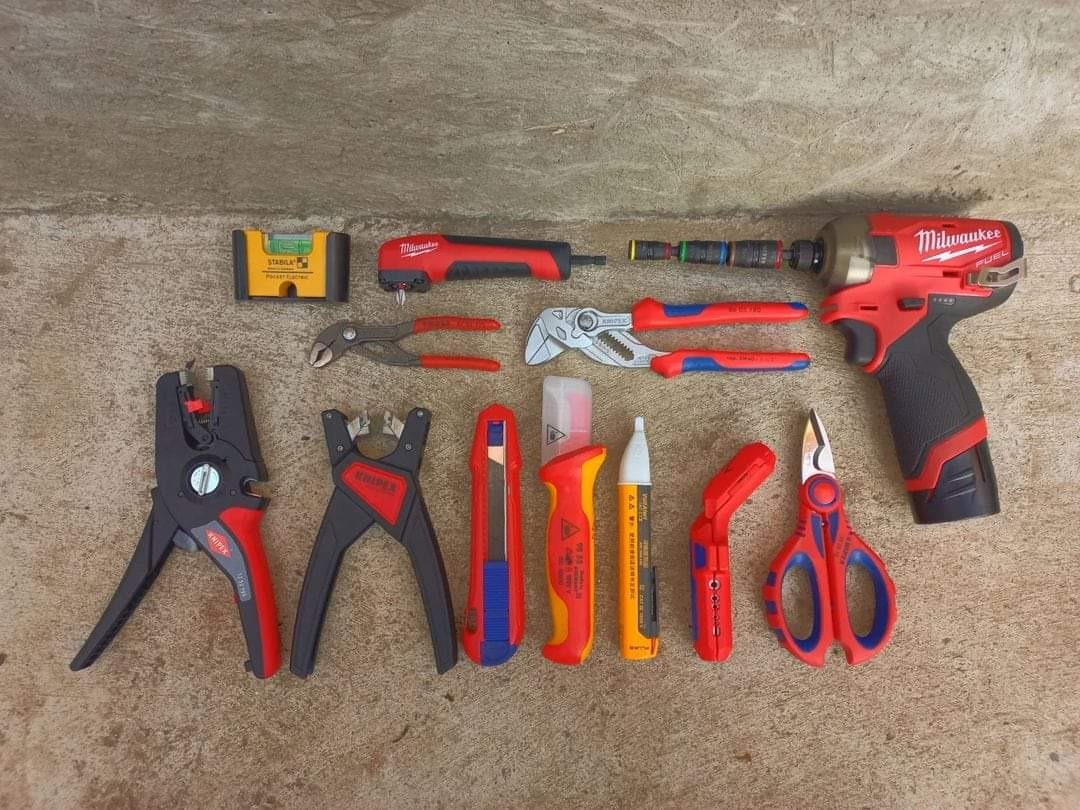 Zestaw narzędzi elektrycznych na szarej powierzchni: wkrętarka udarowa Milwaukee z wymiennymi bitami, poziomica Stabila, szczypce, noże, tester napięcia oraz nożyczki do kabli.