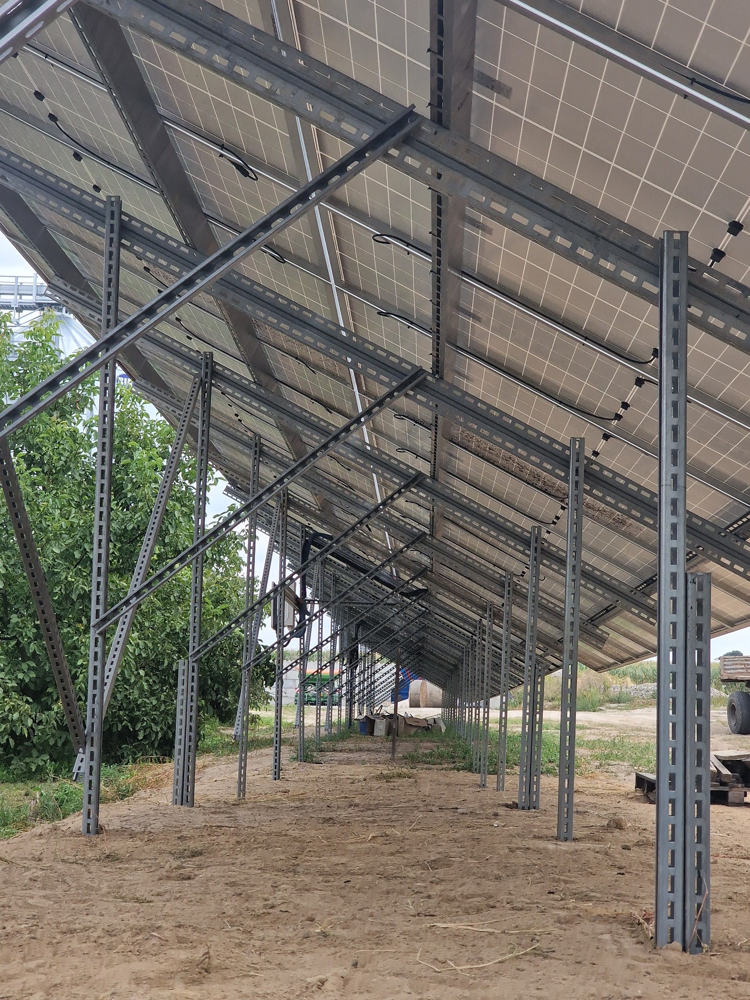 Konstrukcja wsporcza pod panele słoneczne w trakcie montażu, widoczne stalowe profile i częściowo zamontowane panele, tło stanowi grunt i zieleń.