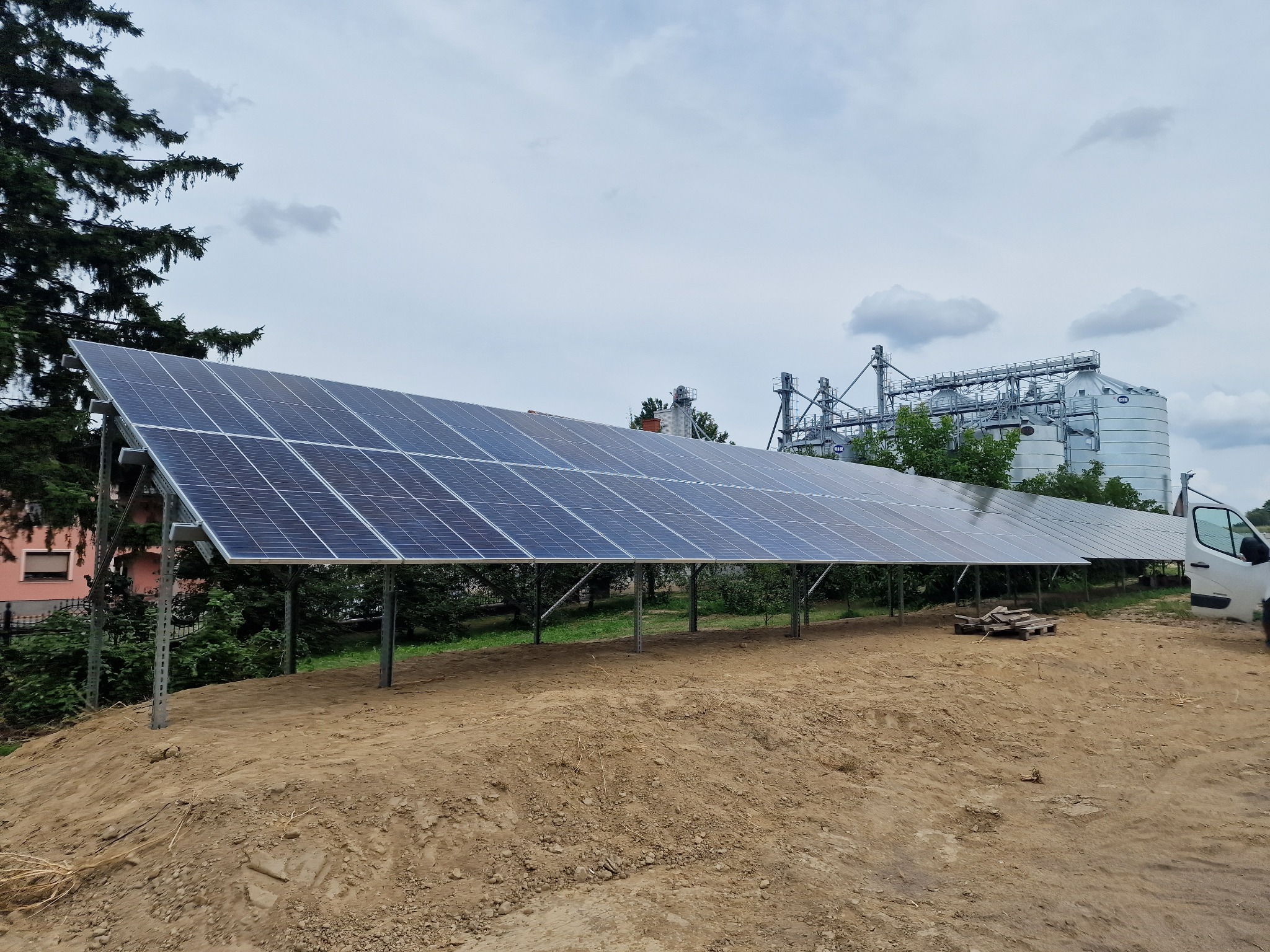 Długi rząd paneli fotowoltaicznych na metalowej konstrukcji, zainstalowany na piaszczystym terenie z widokiem na budynki gospodarcze i częściowo widoczny samochód dostawczy.