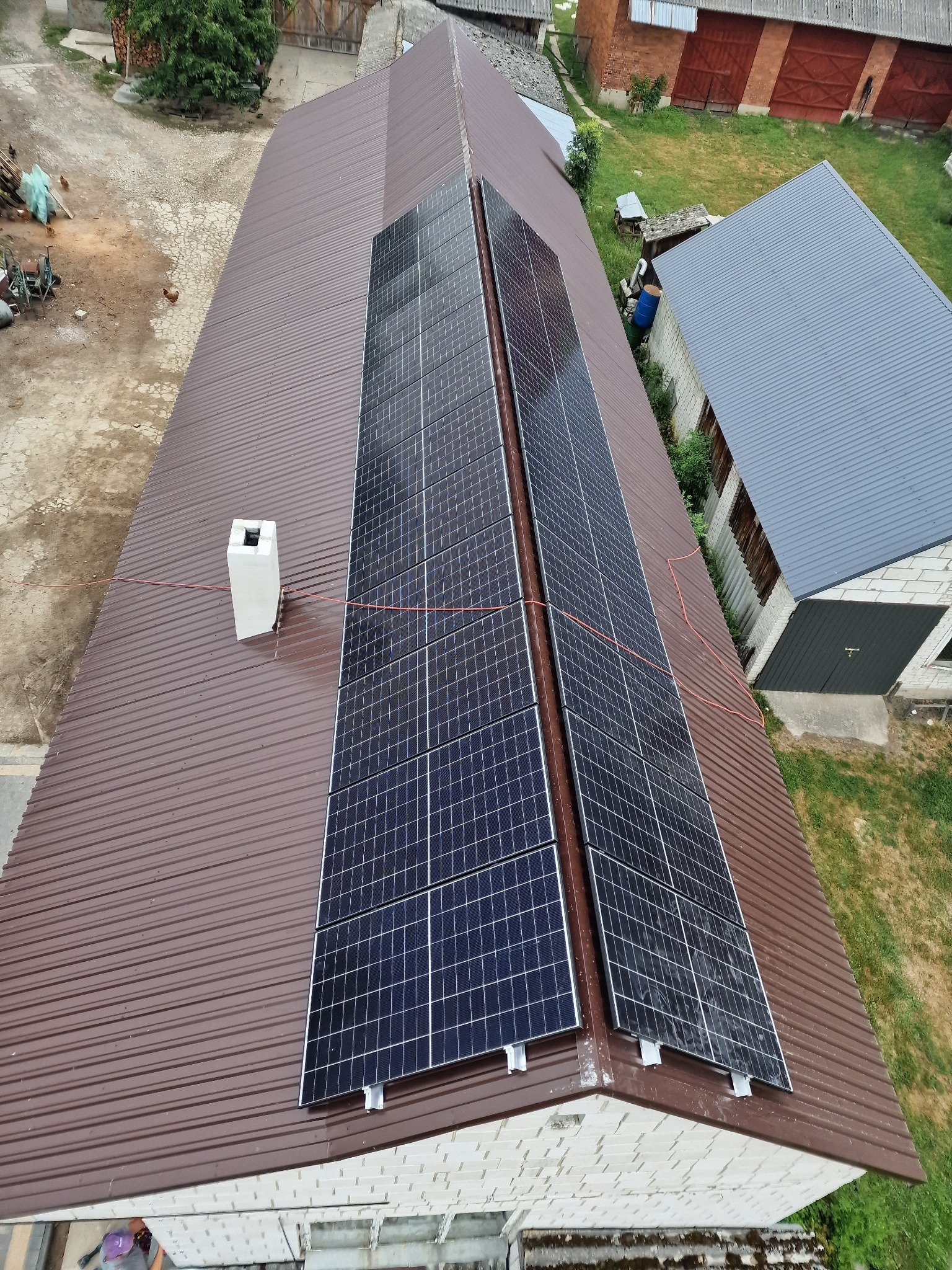 Widok z góry na brązowy dach z blachy trapezowej z dwoma rzędami paneli fotowoltaicznych; widoczny biały komin i przewody elektryczne. W tle zabudowania gospodarcze i zieleń.