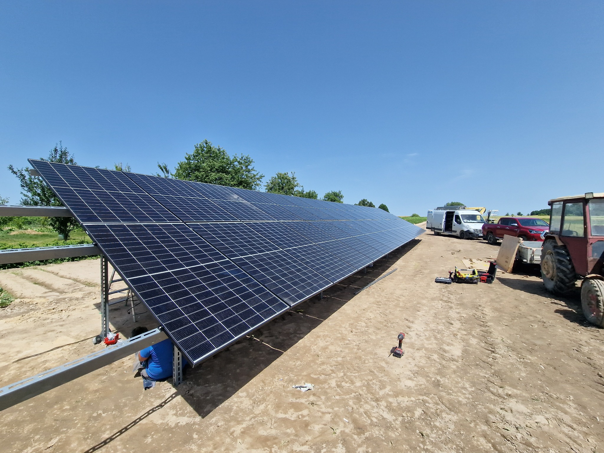 Montaż paneli słonecznych na konstrukcji stalowej w terenie wiejskim, widoczny pracownik w niebieskiej koszulce, samochody dostawcze i ciągnik w tle.