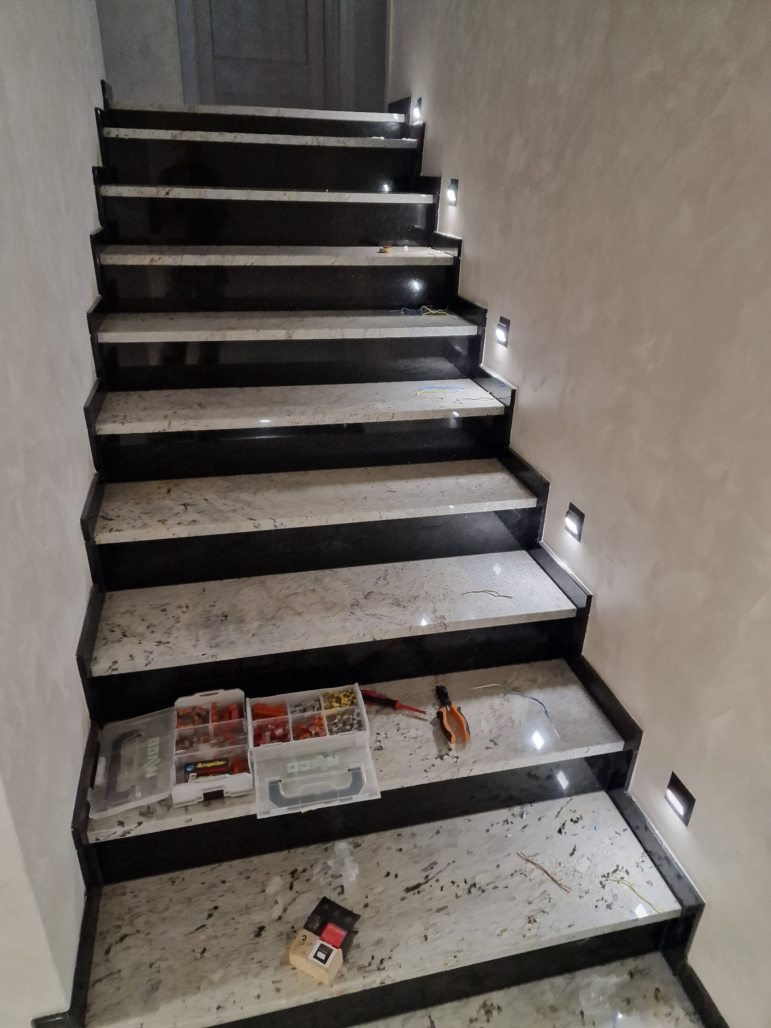 Montaż oświetlenia schodowego: widok na marmurowe schody z zamontowanymi, małymi lampkami LED w ścianie, skrzynki z elementami elektrycznymi i narzędzia na stopniach.