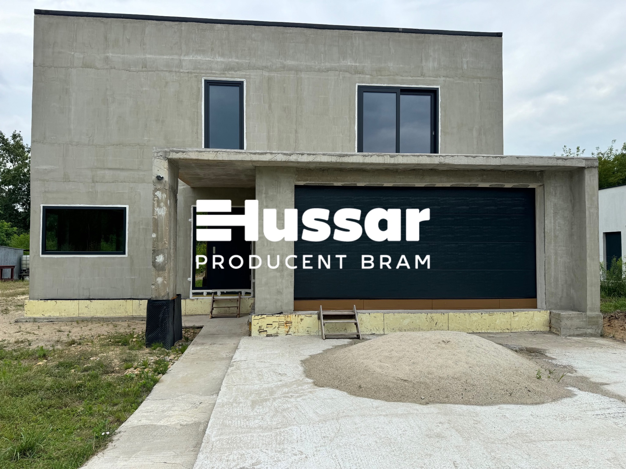 Surowy stan domu jednorodzinnego z zamontowaną czarną bramą garażową i logo producenta na bramie, widoczny stos piasku przed wjazdem do garażu.