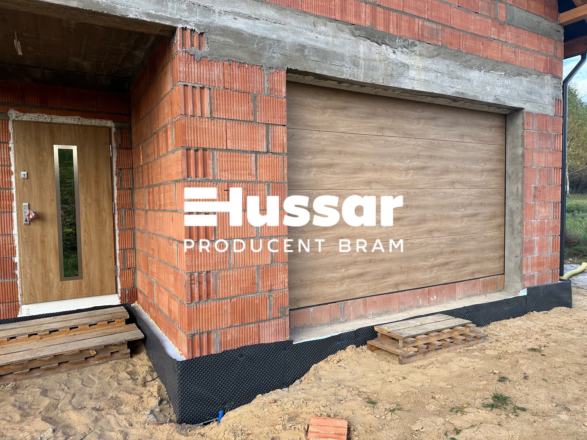 Brama garażowa segmentowa z drewnopodobnym panelem, zamontowana w surowym stanie budynku z czerwonej cegły, widoczne logo producenta, teren wokół w trakcie prac wykończeniowych.