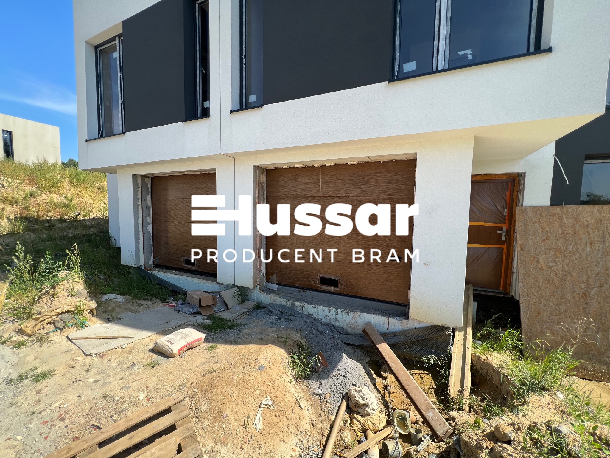 Nowoczesny dom z dwiema bramami garażowymi w trakcie montażu, widoczny bałagan na placu budowy i logo producenta bram na bramach.