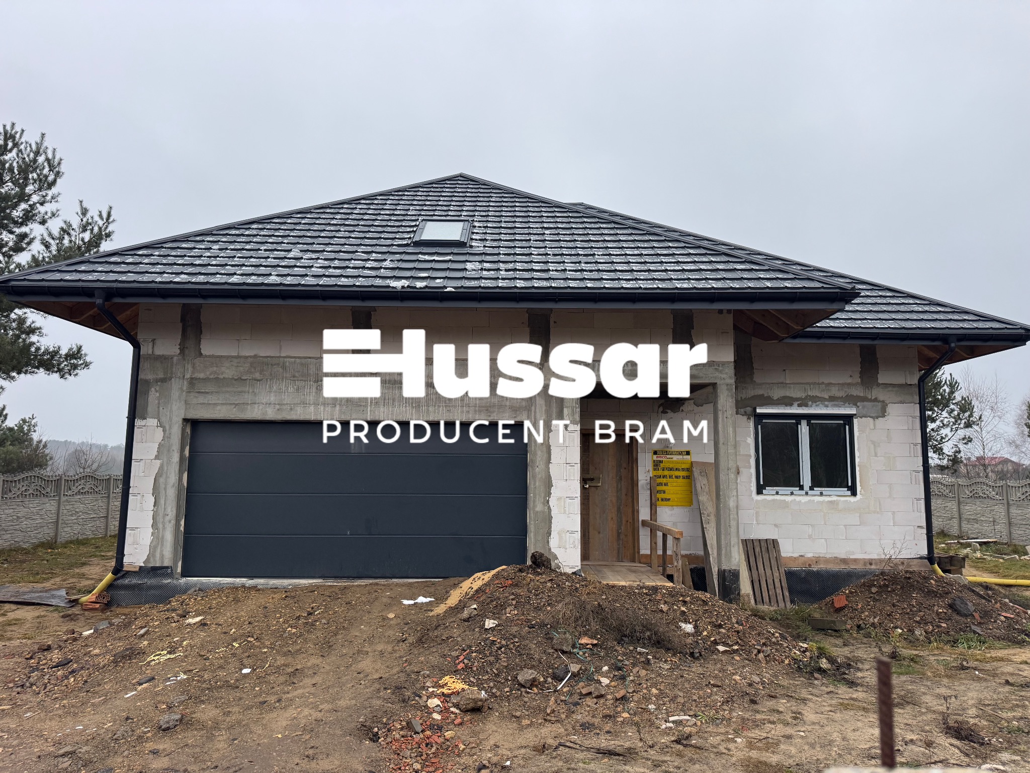 Szara brama garażowa segmentowa w nowo budowanym domu z białego pustaka, z widocznym logo producenta na elewacji i niezagospodarowanym terenem przed budynkiem.