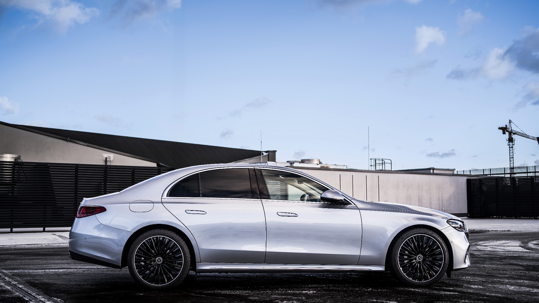 Srebrny samochód osobowy marki Mercedes-Benz klasy S, widok z boku na tle budynku z czarnym dachem i żurawiem budowlanym w oddali.