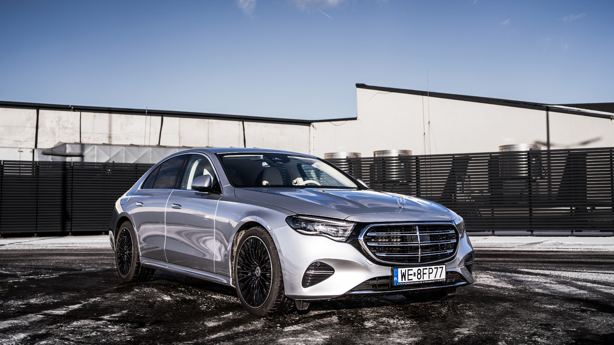 Srebrny samochód osobowy marki Mercedes z czarnymi felgami, zaparkowany na parkingu z widocznym budynkiem w tle.