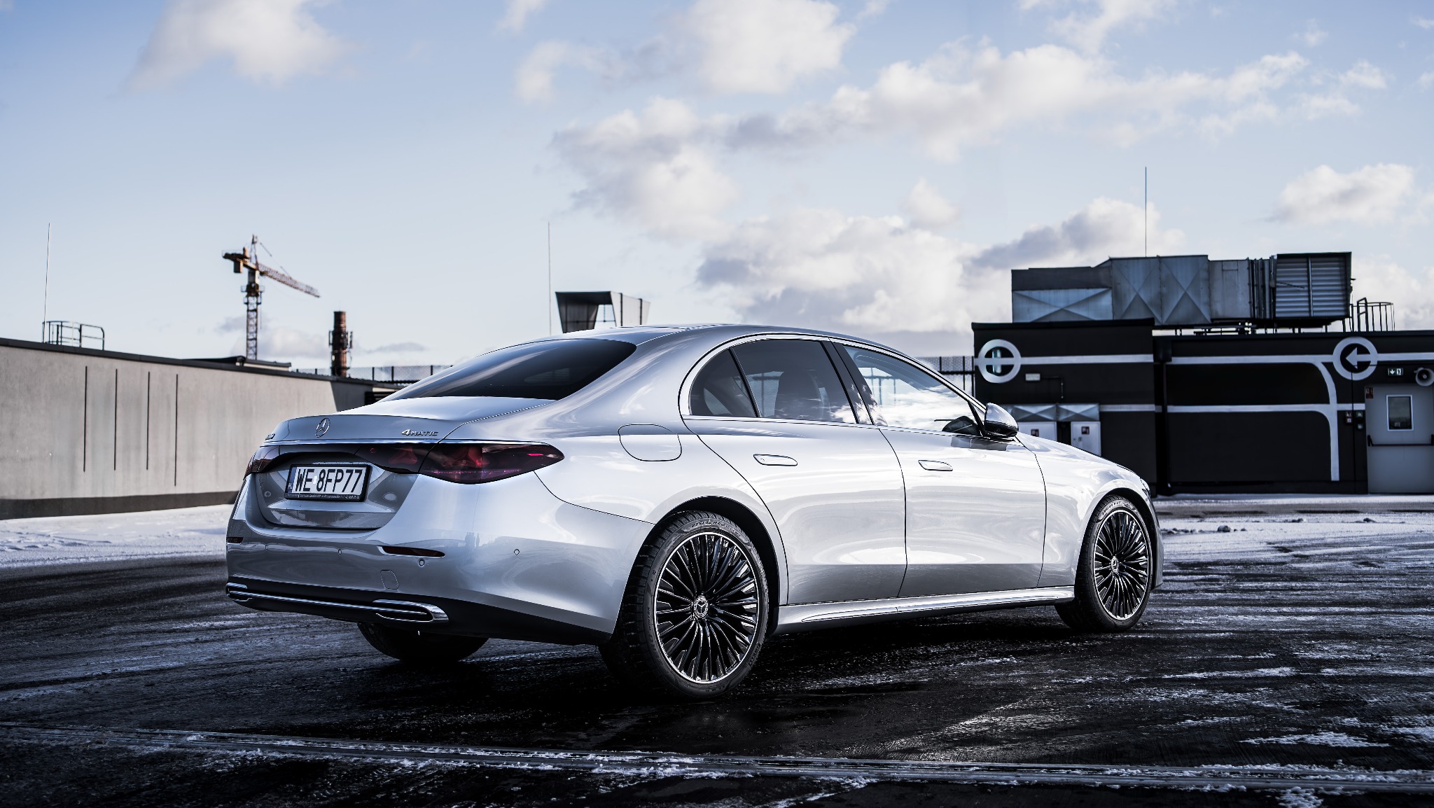 Srebrny sedan Mercedes-Benz S-Class 4Matic, widziany z tyłu, na tle dachu budynku z konstrukcją wentylacyjną i żurawiem budowlanym w tle, w pochmurny dzień.