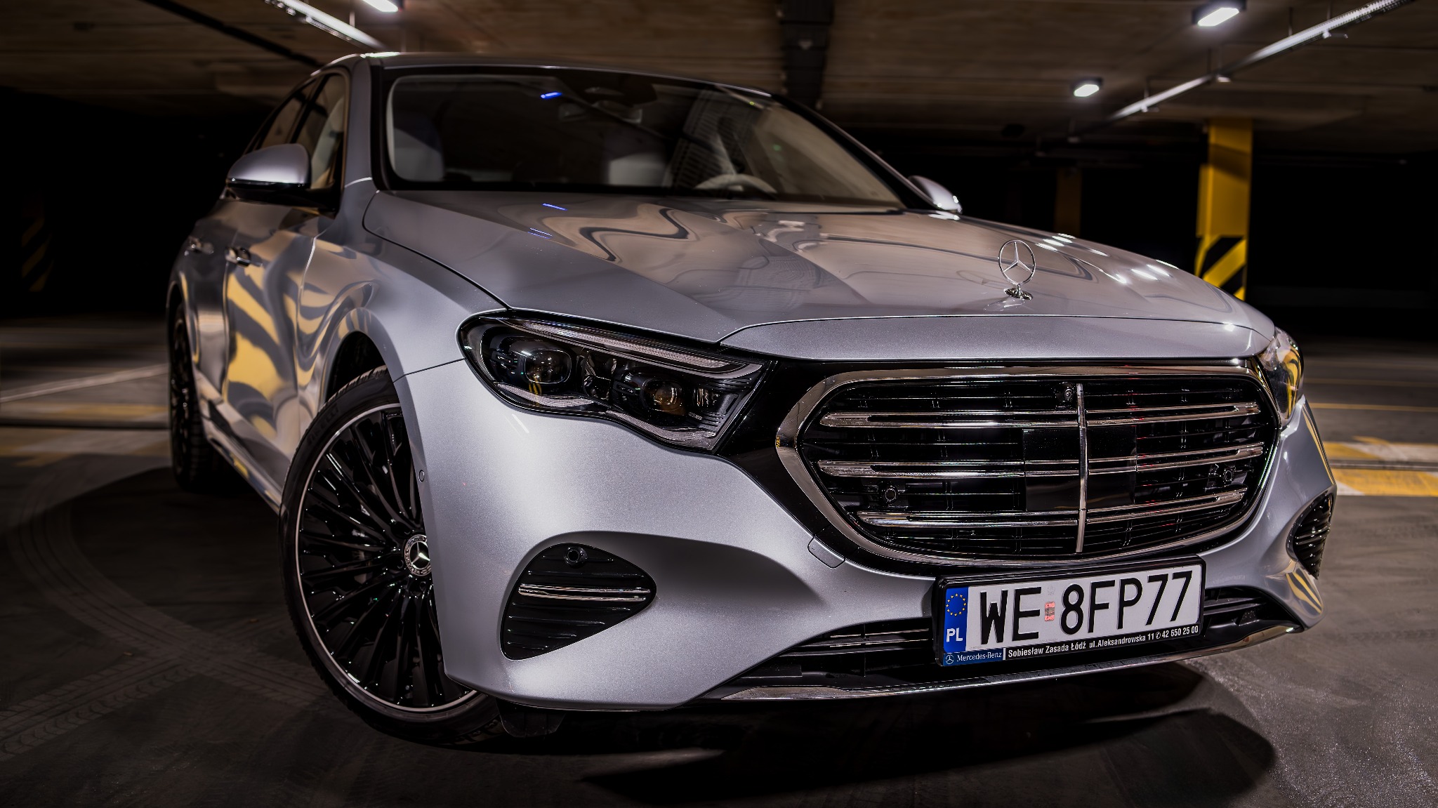 Srebrny Mercedes-Benz z czarnymi felgami, sfotografowany pod kątem w garażu podziemnym, widoczne detale grilla i reflektorów.