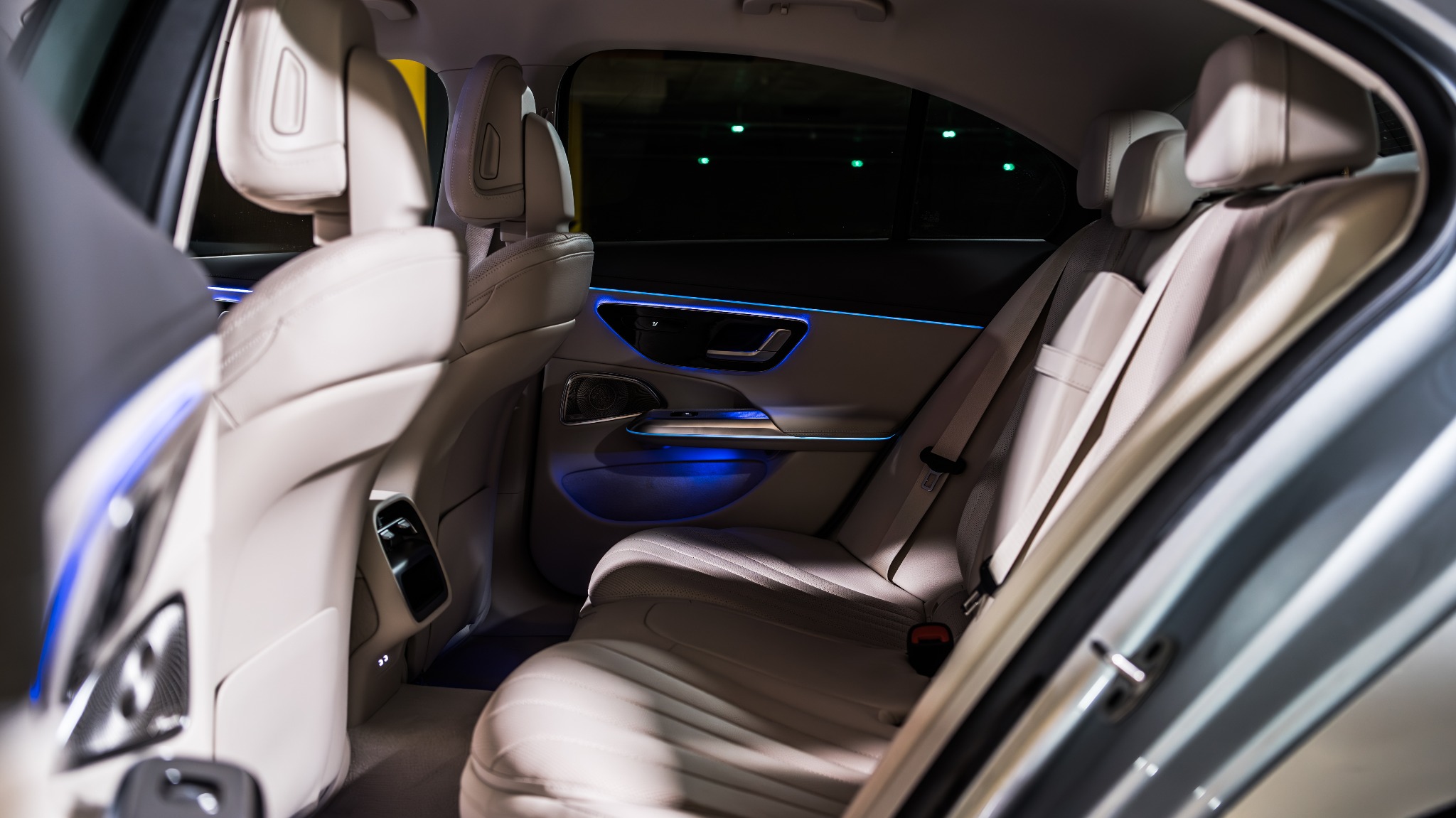 Wnętrze samochodu z beżową skórzaną tapicerką i niebieskim oświetleniem ambientowym, widok na tylne siedzenia z perspektywy pasażera z przodu.