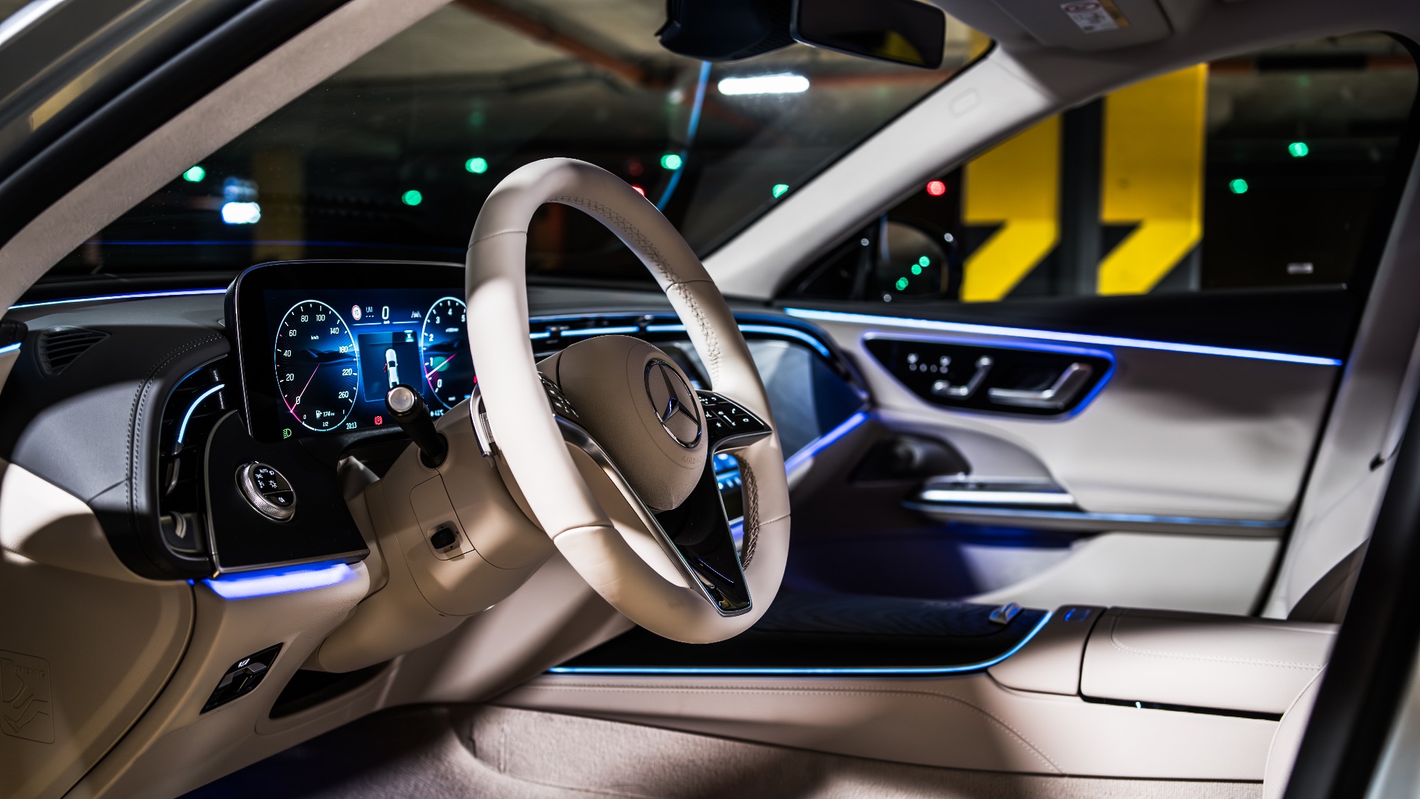 Wnętrze luksusowego samochodu Mercedes z beżową skórzaną tapicerką, podświetleniem LED i cyfrowym kokpitem, widok z perspektywy kierowcy na kierownicę z logo.