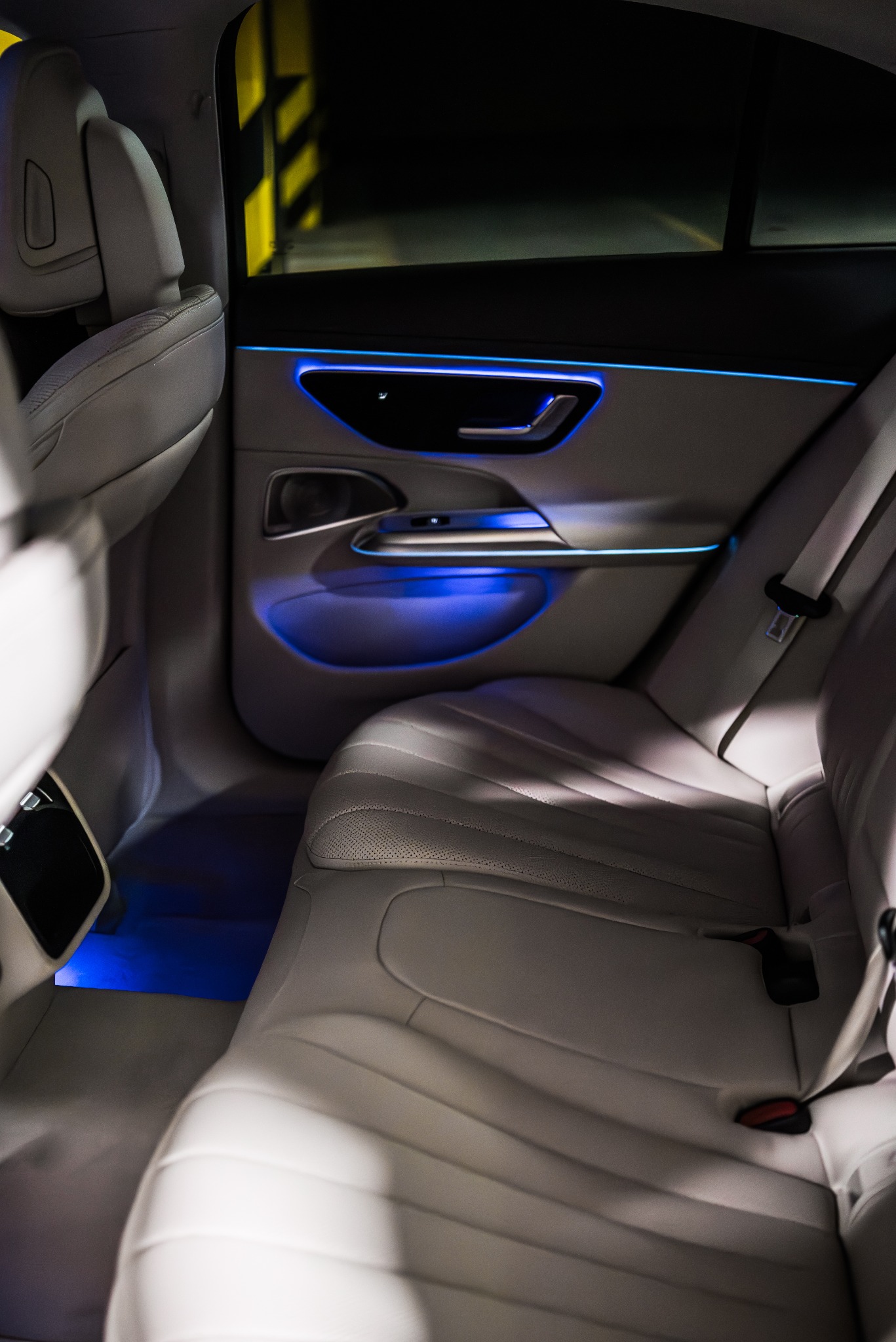 Wnętrze luksusowego samochodu z jasną skórzaną tapicerką, podświetleniem LED w kolorze niebieskim i widocznymi tylnymi siedzeniami oraz fragmentem słupka z żółto-czarnymi pasami w tle.