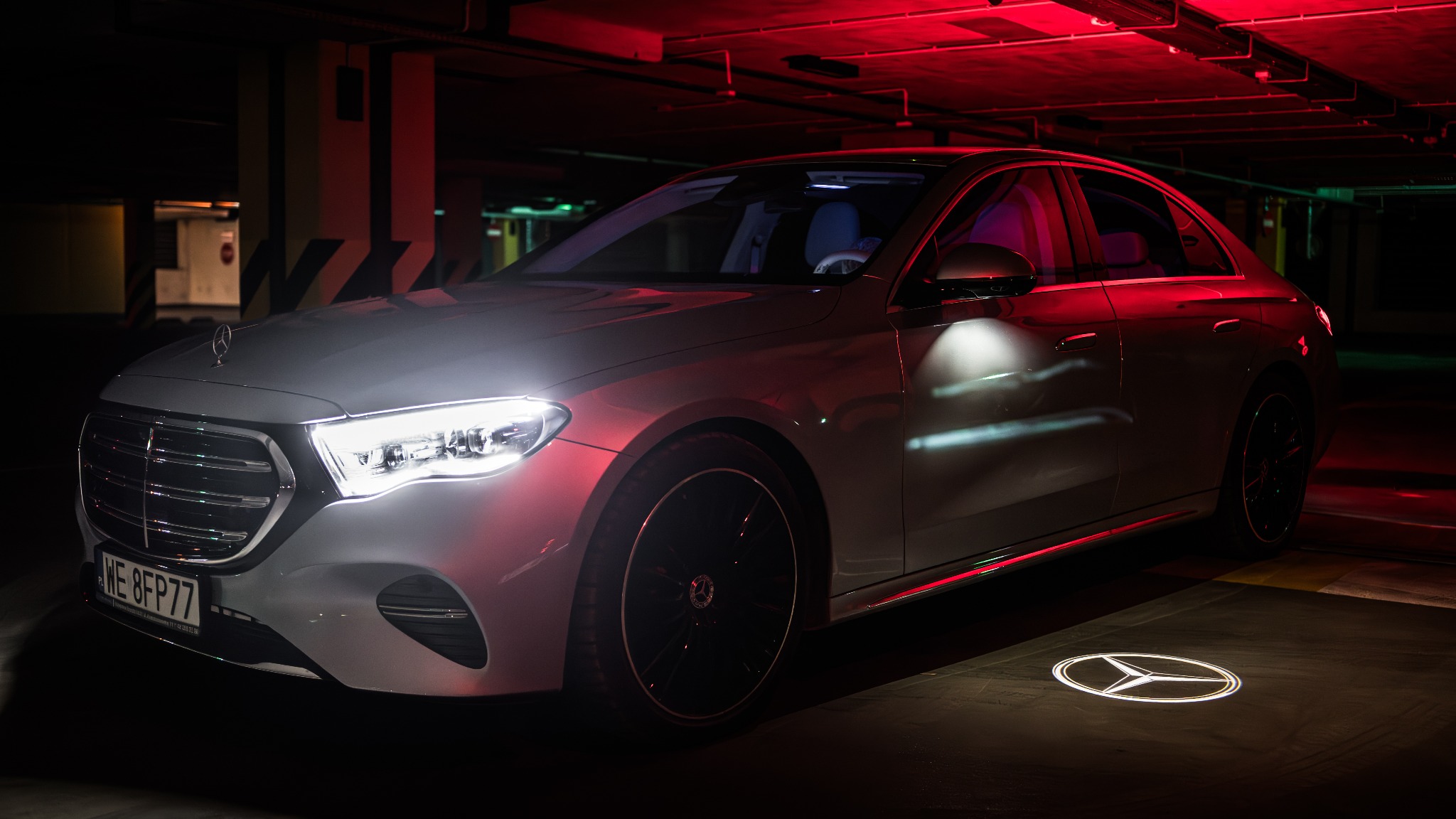 Srebrny Mercedes-Benz w garażu podziemnym w Ożarowie Mazowieckim, z podświetlanym logo na podłożu i czerwonym oświetleniem sufitowym.