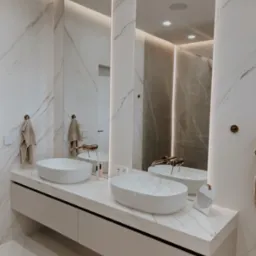 Nowoczesna łazienka z marmurowymi ścianami i blatem, dwie owalne umywalki, złota armatura i podświetlane lustra. Minimalistyczny design, eleganckie wykończenie.