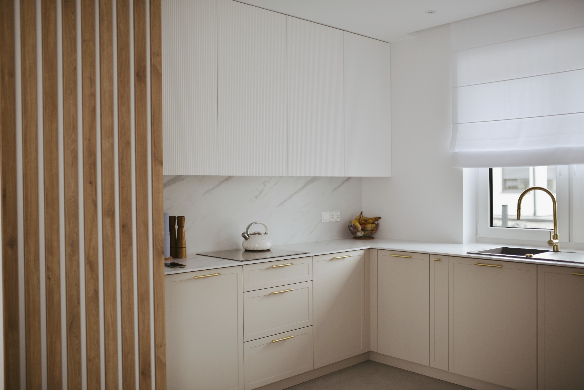 Jasna kuchnia z frezowanymi frontami szafek, złotymi uchwytami i drewnianą ażurową ścianką działową, widok na czajnik na płycie grzewczej i owoce w ozdobnej misie.