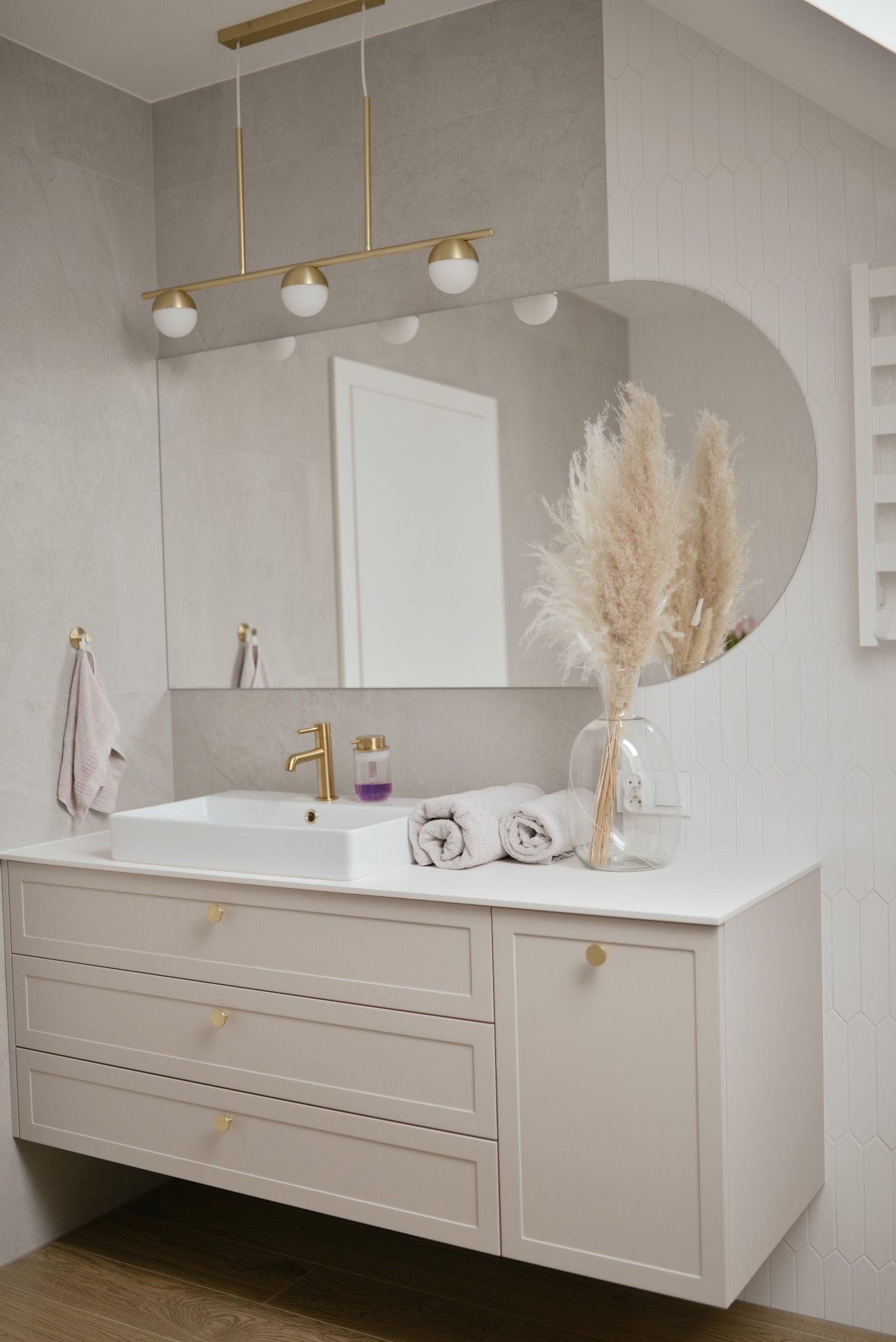 Nowoczesna łazienka z beżową szafką pod umywalką, złotą armaturą i okrągłym lustrem, dekorowana trawą pampasową w szklanym wazonie.