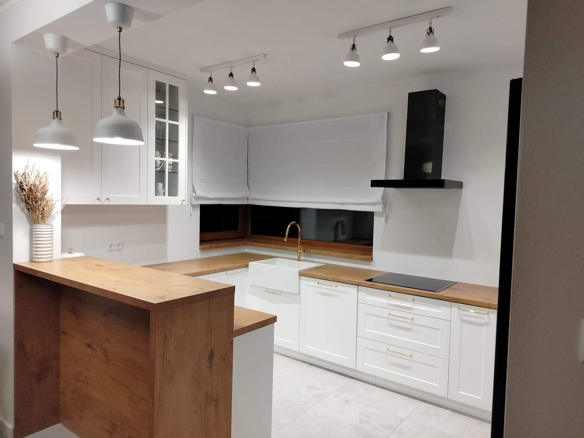 Jasna kuchnia z białymi szafkami, drewnianym blatem i złotą armaturą, oświetlona wiszącymi lampami i reflektorami sufitowymi, z widocznym oknem z roletą rzymską.