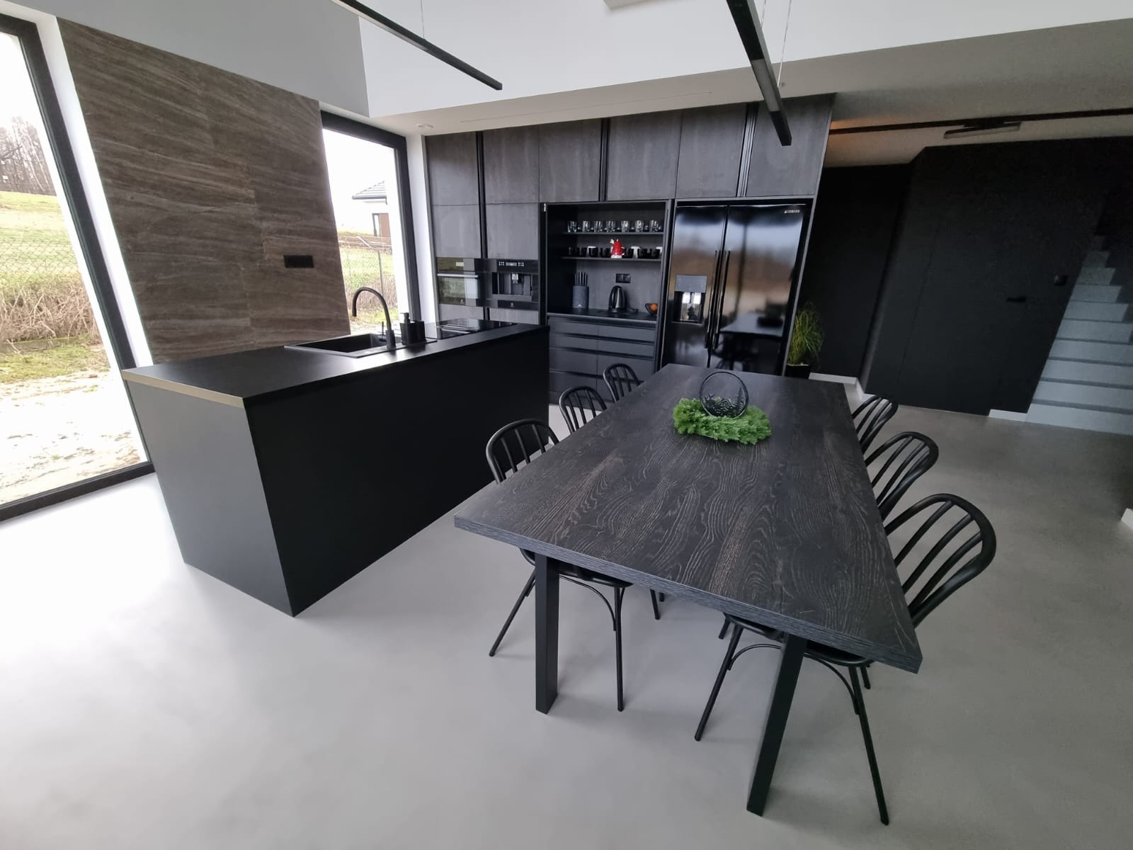 Minimalistyczna, czarna kuchnia z wyspą i dużym oknem, połączona z jadalnią z ciemnym, drewnianym stołem i czarnymi krzesłami. Na stole dekoracja roślinna. Widoczne zabudowane szafki kuchenne...