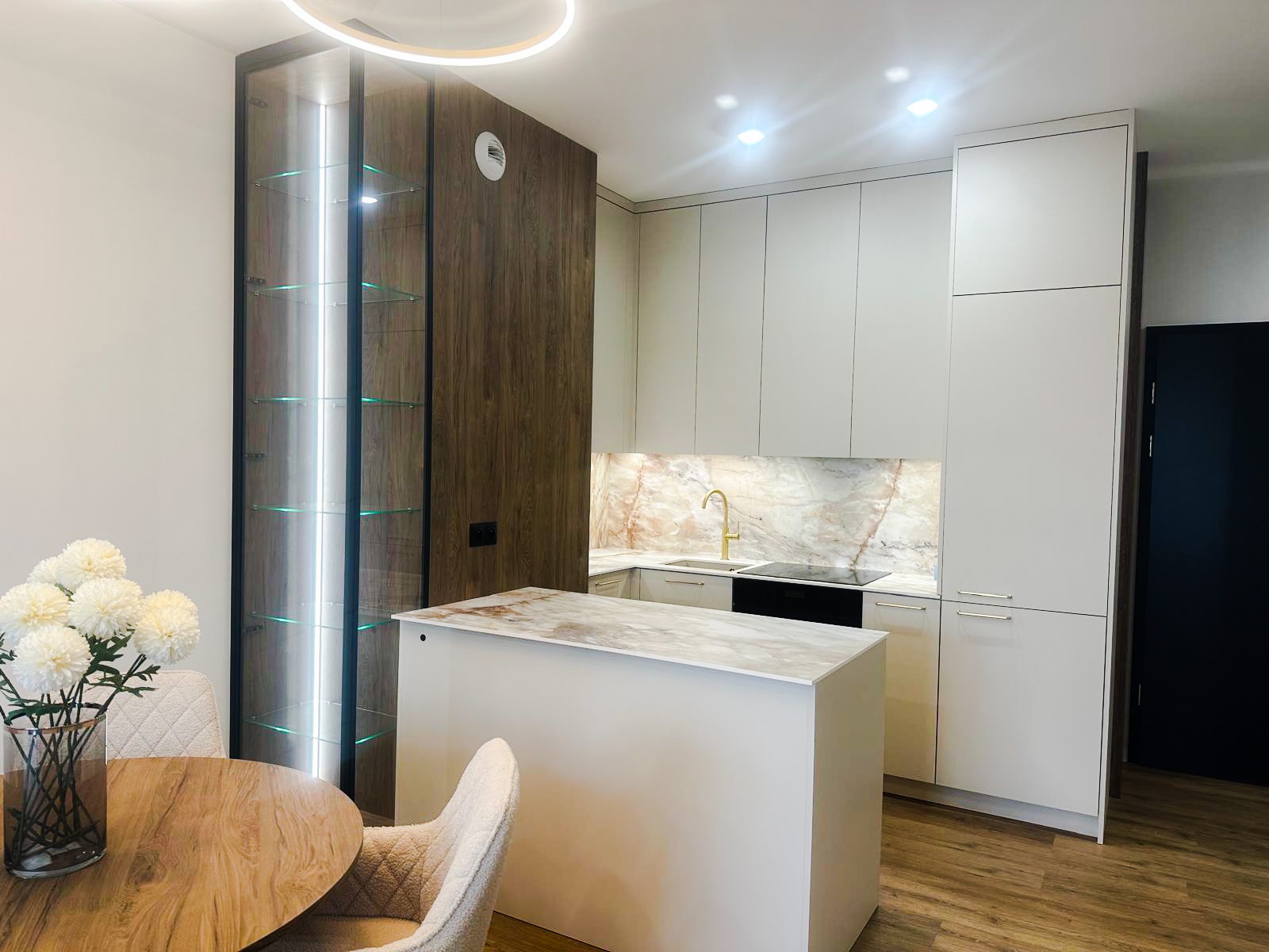 Nowoczesna kuchnia z wyspą, białymi szafkami i marmurowym blatem oraz podświetlana witryna z szklanymi półkami, widok z jadalni z drewnianym stołem i bukietem białych kwiatów.