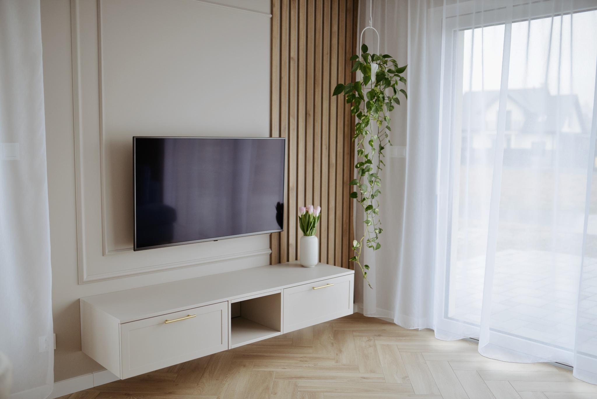 Nowoczesny salon z wiszącą szafką RTV w odcieniach beżu, drewnianą ścianką dekoracyjną, telewizorem i bukietem tulipanów w białym wazonie, uzupełniony zwisającą rośliną doniczkową i jasnymi zasłonami.
