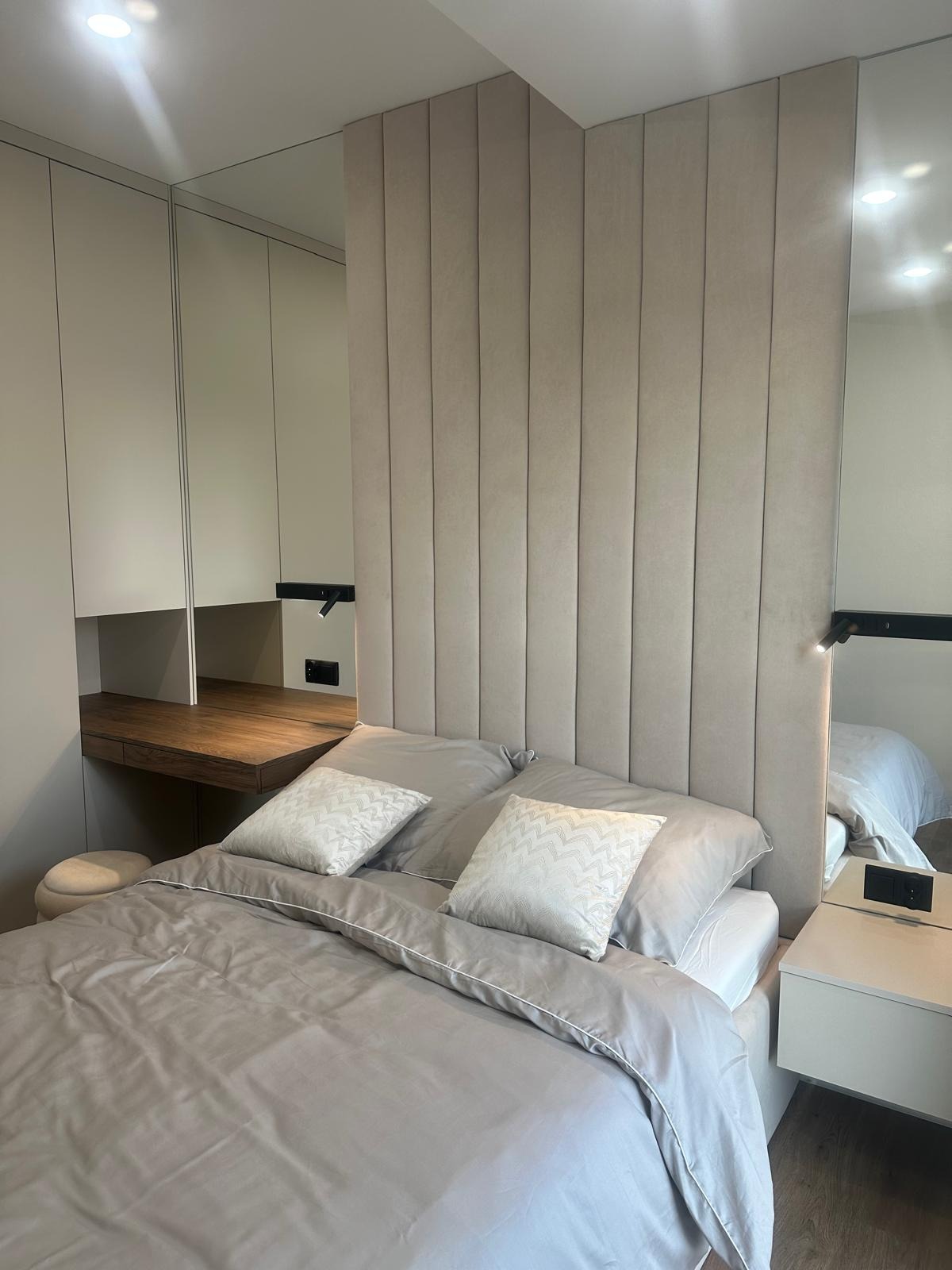 Sypialnia z łóżkiem, tapicerowanym panelem ściennym w odcieniach beżu, biurkiem z drewnianym blatem w zabudowie i lustrem.