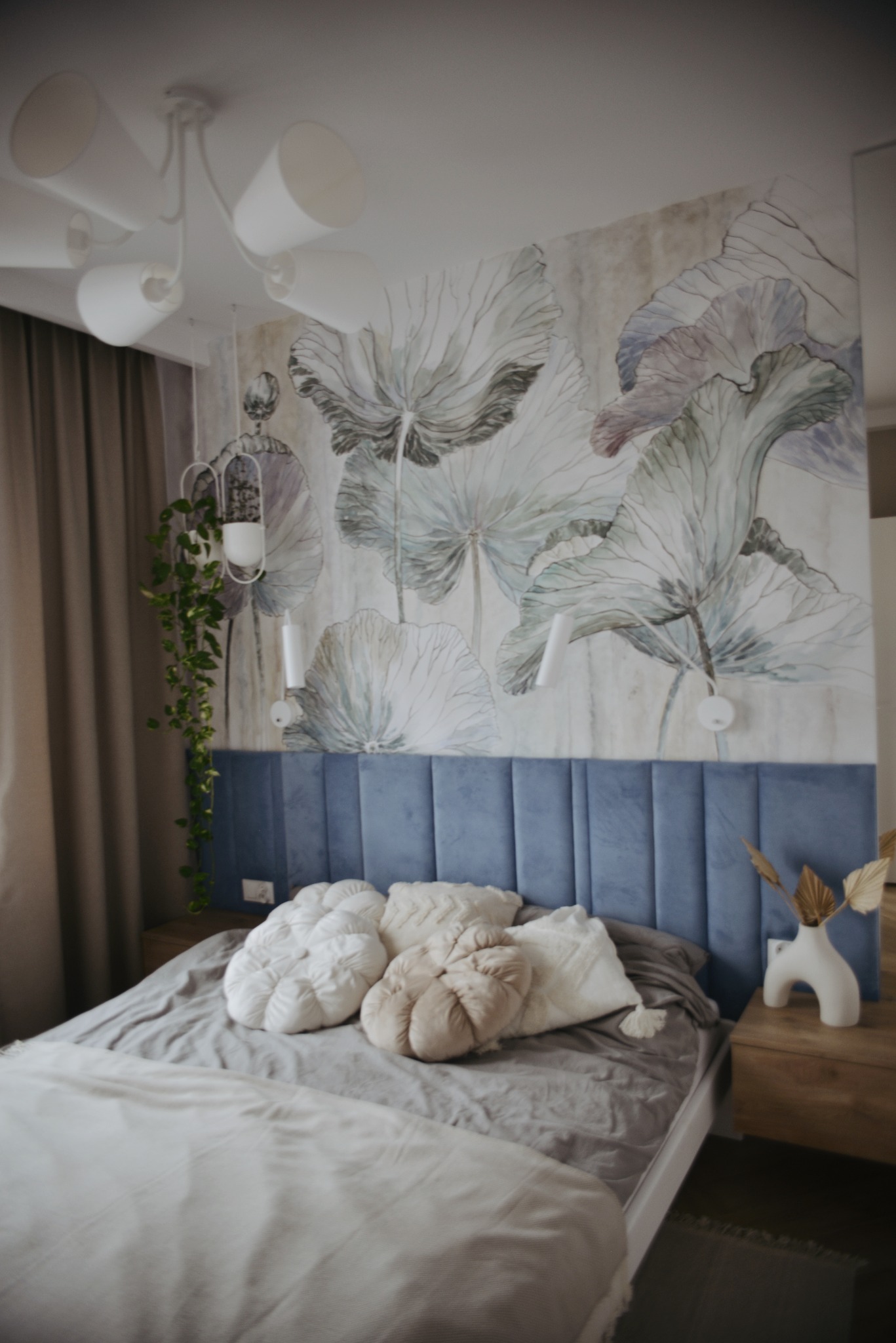 Sypialnia z niebieskim tapicerowanym zagłówkiem, dekoracyjnymi poduszkami w kształcie dyni, lampą wiszącą z białymi kloszami i ścianą z motywem roślinnym w odcieniach szarości i fioletu.