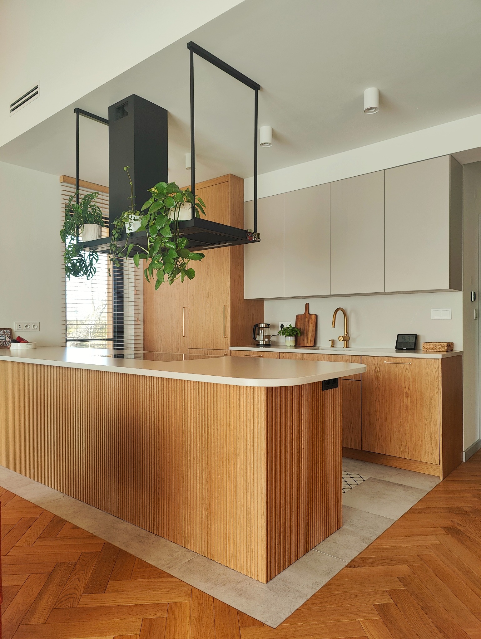 Nowoczesna kuchnia z wyspą wykończoną ryflowanym drewnem, wiszącym okapem z roślinami i parkietem w jodełkę.