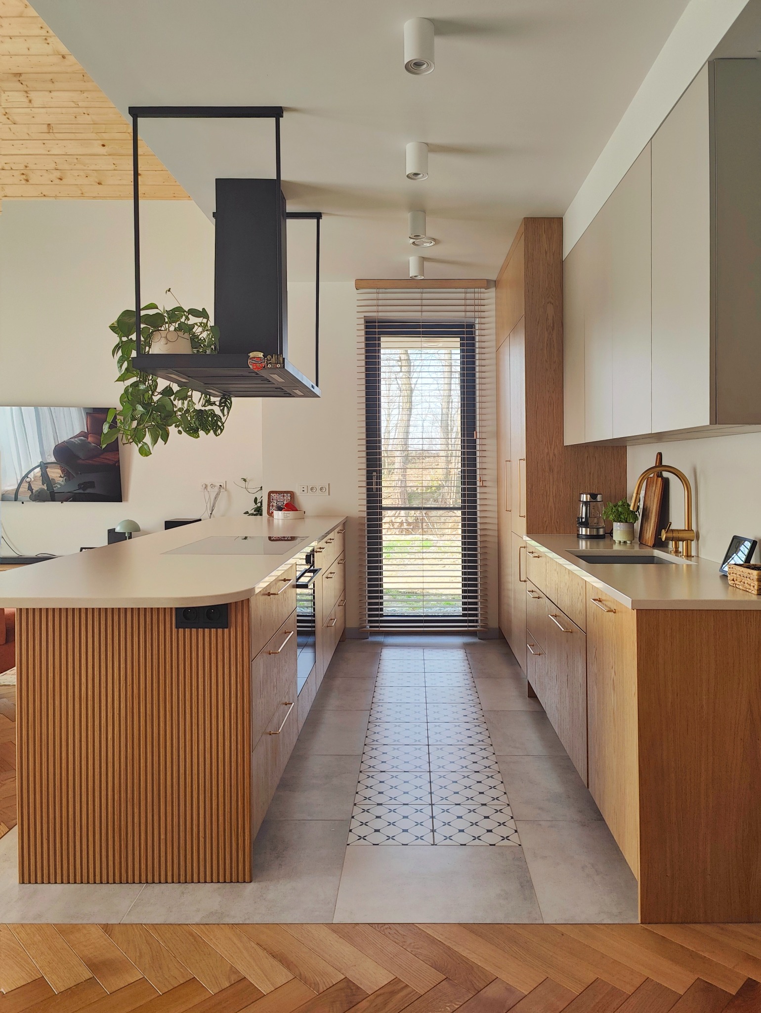 Nowoczesna kuchnia z drewnianymi frontami szafek, blatem w jasnym odcieniu beżu i podłogą wyłożoną płytkami w geometryczny wzór. Na wyspie kuchennej zamontowana płyta indukcyjna. Nad wyspą...