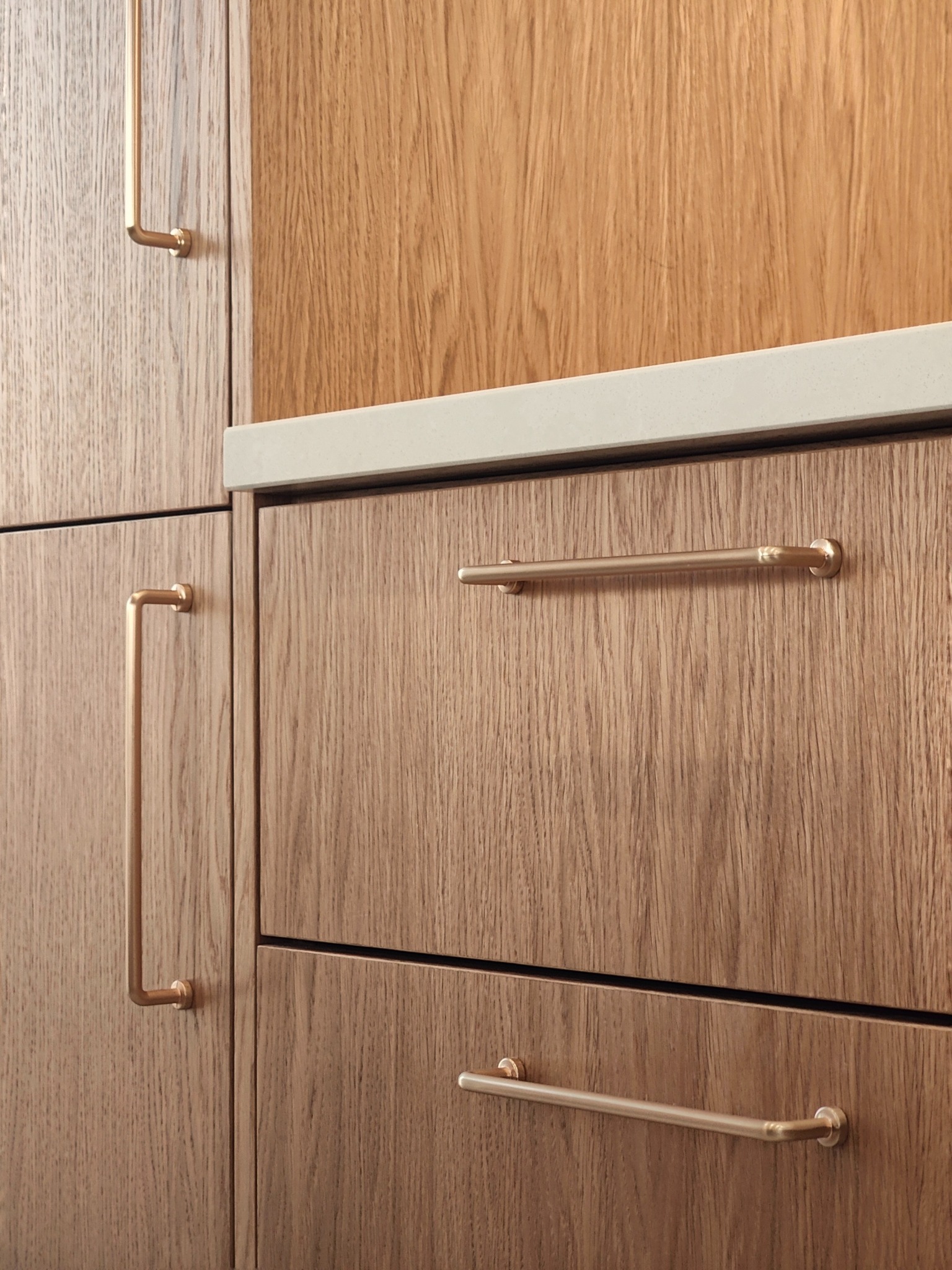 Szczegółowe ujęcie drewnianych frontów szuflad i szafek z poziomymi, metalowymi uchwytami w kolorze mosiądzu, z widocznym blatem o jasnej, jednolitej powierzchni.