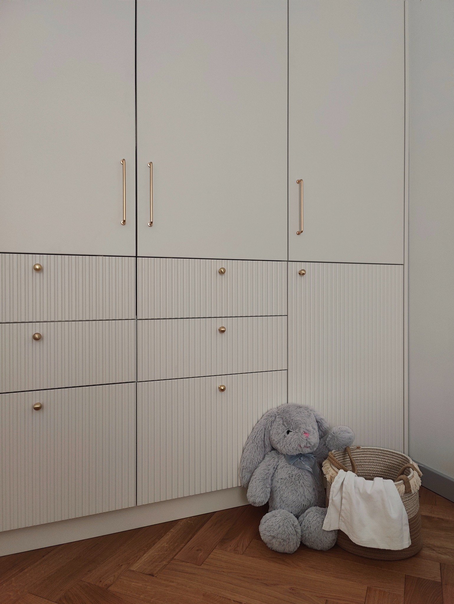 Jasnoszara szafa wnękowa z frezowanymi frontami szuflad i złotymi uchwytami, uzupełniona pluszowym królikiem i koszem z tkaniną na parkiecie w jodełkę.