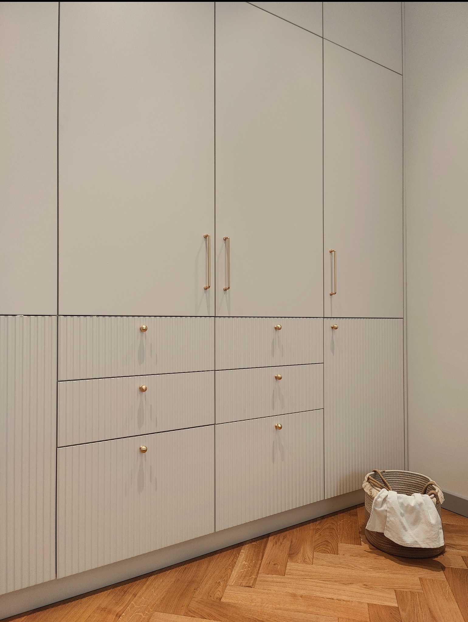Wysoka, zabudowana szafa z szufladami i górnymi szafkami w odcieniach beżu, z ryflowanymi frontami szuflad i złotymi uchwytami, na parkiecie ułożonym w jodełkę, z koszem na pranie w rogu.