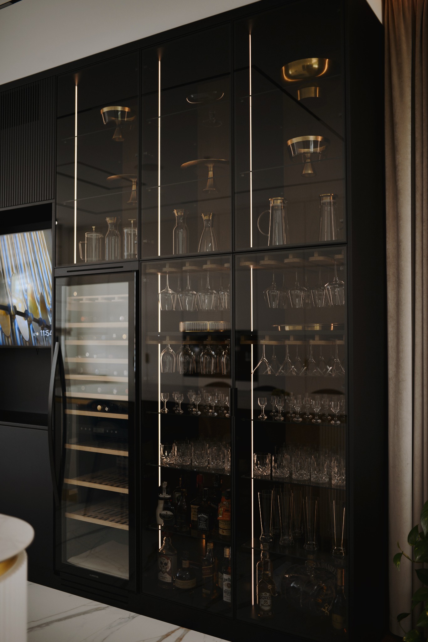 Czarna zabudowa meblowa z przeszklonymi witrynami, podświetlana LED, eksponująca szkło i alkohol, z wbudowaną chłodziarką do wina. Elegancki design.