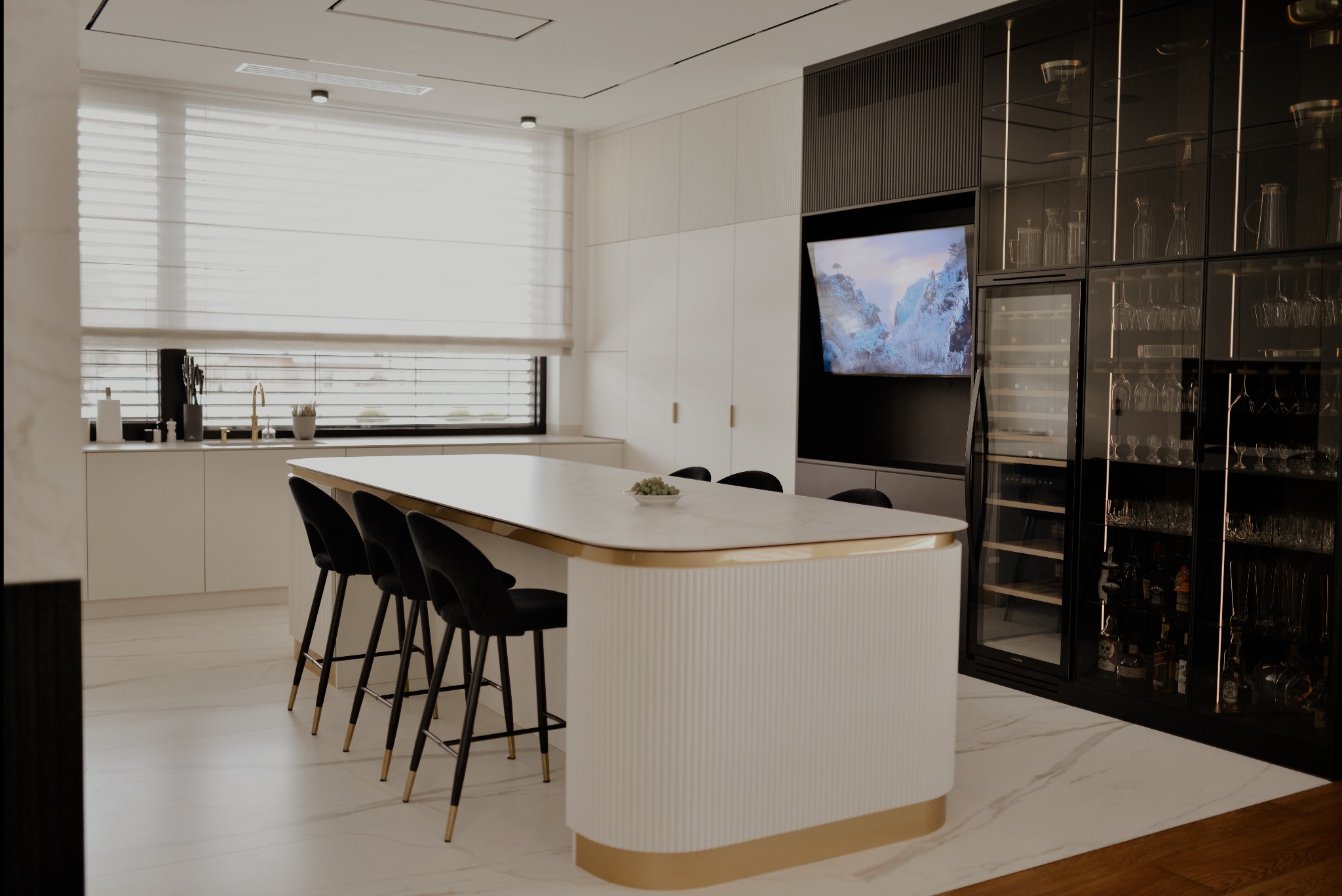 Elegancka kuchnia z wyspą, czarnymi hokerami i zabudową z barkiem. Białe fronty szafek, marmurowy blat, złote akcenty. Nowoczesny design z dbałością o detale.