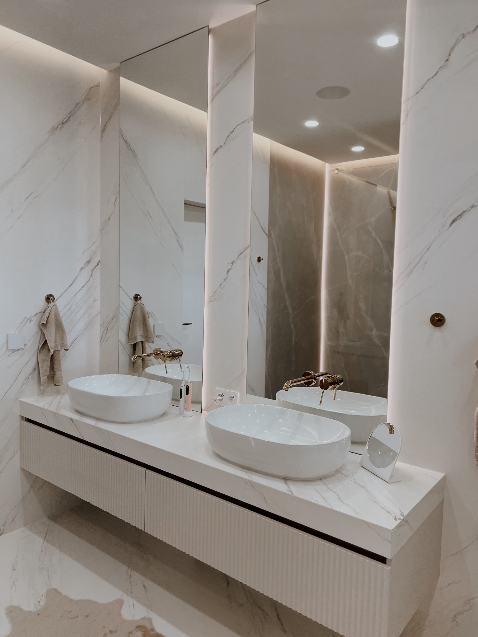 Nowoczesna łazienka z marmurowymi ścianami i blatem, dwie owalne umywalki, złota armatura i podświetlane lustra. Minimalistyczny design, eleganckie wykończenie.