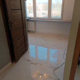 Sebastian Martirosyan - Nowo ułożona, błyszcząca podłoga z płytek imitujących marmur w jasnym pomieszczeniu z oknem i grzejnikiem. Widoczna otwarta, drewniana rama drzwi i narzędzia do układania płytek przy podłodze.