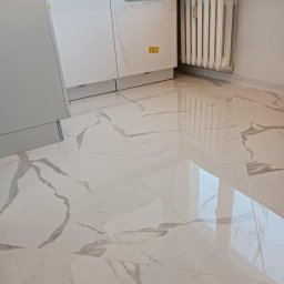 Sebastian Martirosyan - Błyszczące, białe płytki podłogowe imitujące marmur, ułożone w nowym pomieszczeniu z grzejnikiem i częściowo widocznymi białymi szafkami kuchennymi z folią ochronną.