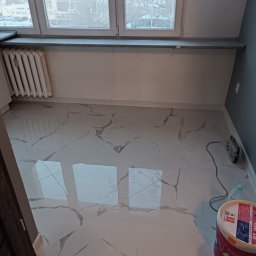 Sebastian Martirosyan - Świeżo położone, błyszczące płytki podłogowe imitujące marmur w pokoju z grzejnikiem pod oknem, widoczny pojemnik z gotową gładzią polimerową oraz radio budowlane.