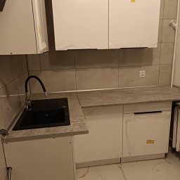 Sebastian Martirosyan - Nowoczesna, minimalistyczna kuchnia w trakcie wykańczania, z białymi szafkami z folią ochronną, czarnym zlewem z elastyczną wylewką i szarymi płytkami na ścianie, widoczne gniazdko elektryczne.