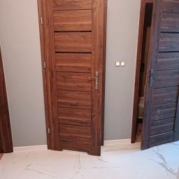 Sebastian Martirosyan - Dwa drewniane drzwi wewnętrzne z poziomymi wstawkami, jeden zamknięty, drugi otwarty, na tle szarej ściany i białej podłogi z marmurowym wzorem.