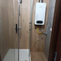 Kabina prysznicowa z zamontowanym piecem gazowym Junkers na ścianie wyłożonej płytkami imitującymi drewno, z ż&oacute;łtą instalacją gazową.