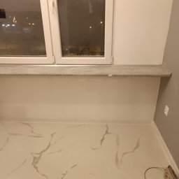 Sebastian Martirosyan - Wykończona wnęka okienna z parapetem z płytek imitujących marmur, białe ściany i podłoga z płytek w podobnym stylu, widoczny fragment szarej ściany oraz narzędzie pomiarowe przy podłodze.